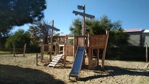 Aire de jeux en bois avec thème pirate, équipée de toboggans et de structures d'escalade, installée sur un sol sablonneux.